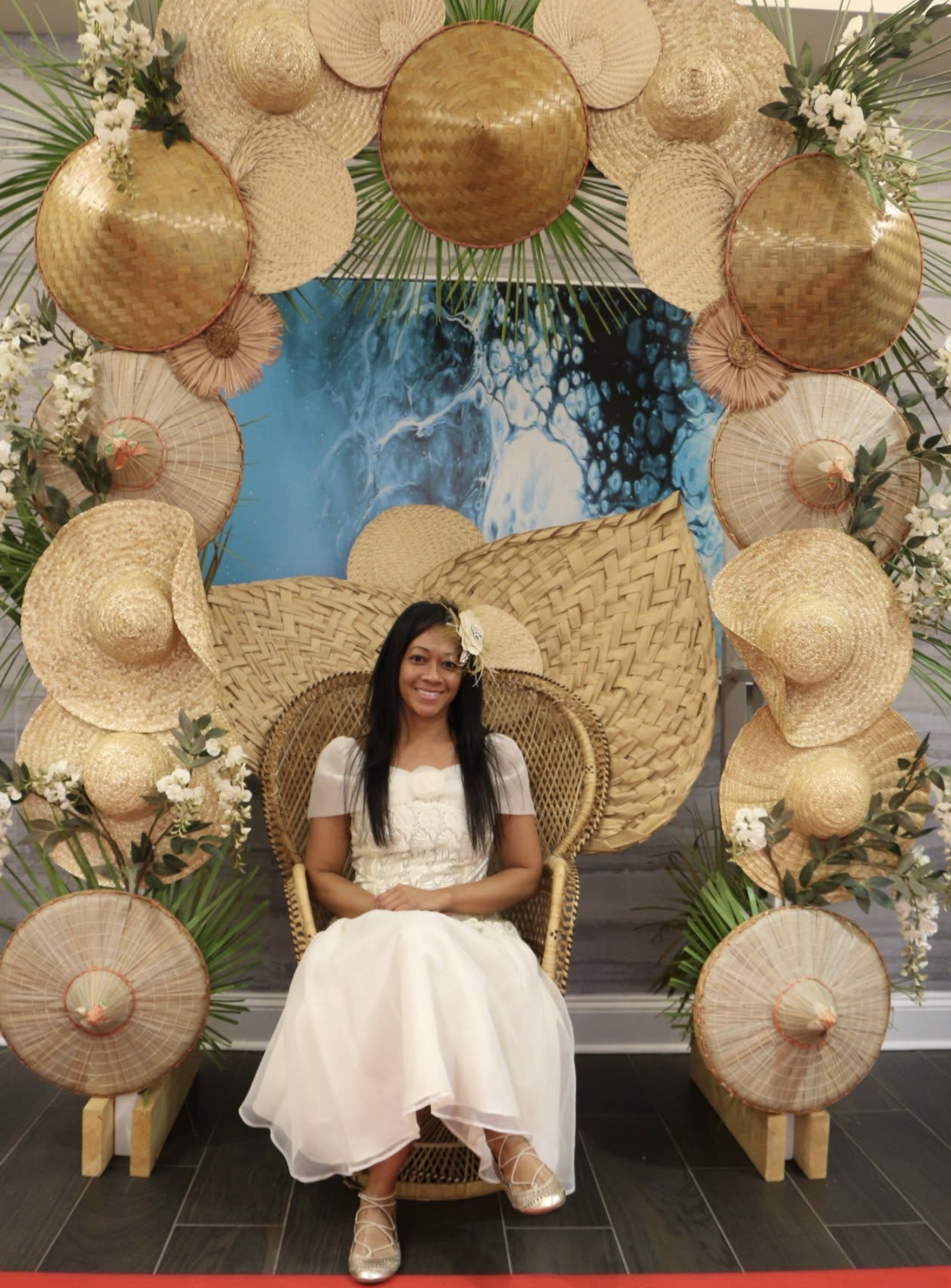 A woman in a white dress sits on a wicker chair in front of a decorative backdrop of straw hats and green foliage, with a blue abstract painting behind her.