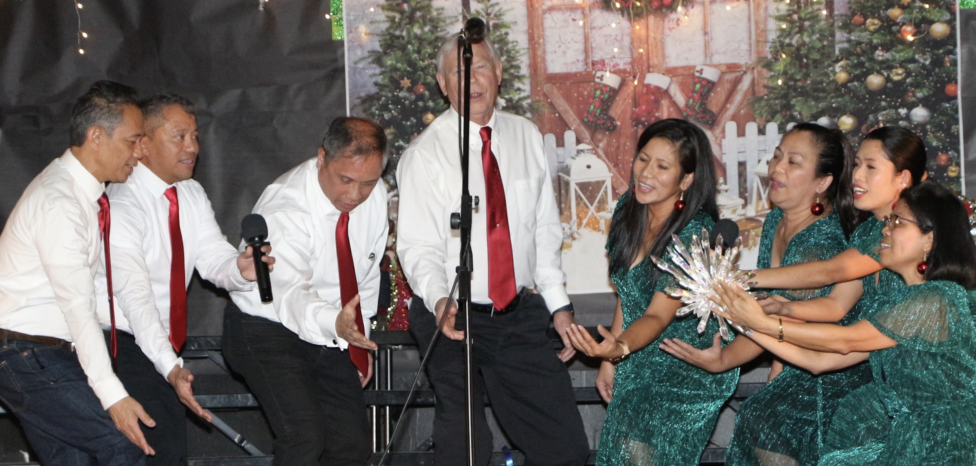 A group of people performing Christmas carol singing on stage with Christmas decorations in the background.