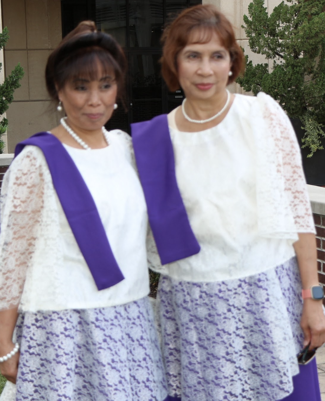 Two women standing outdoors, dressed in white lace tops with purple sashes, wearing pearl jewelry, with greenery in the background.