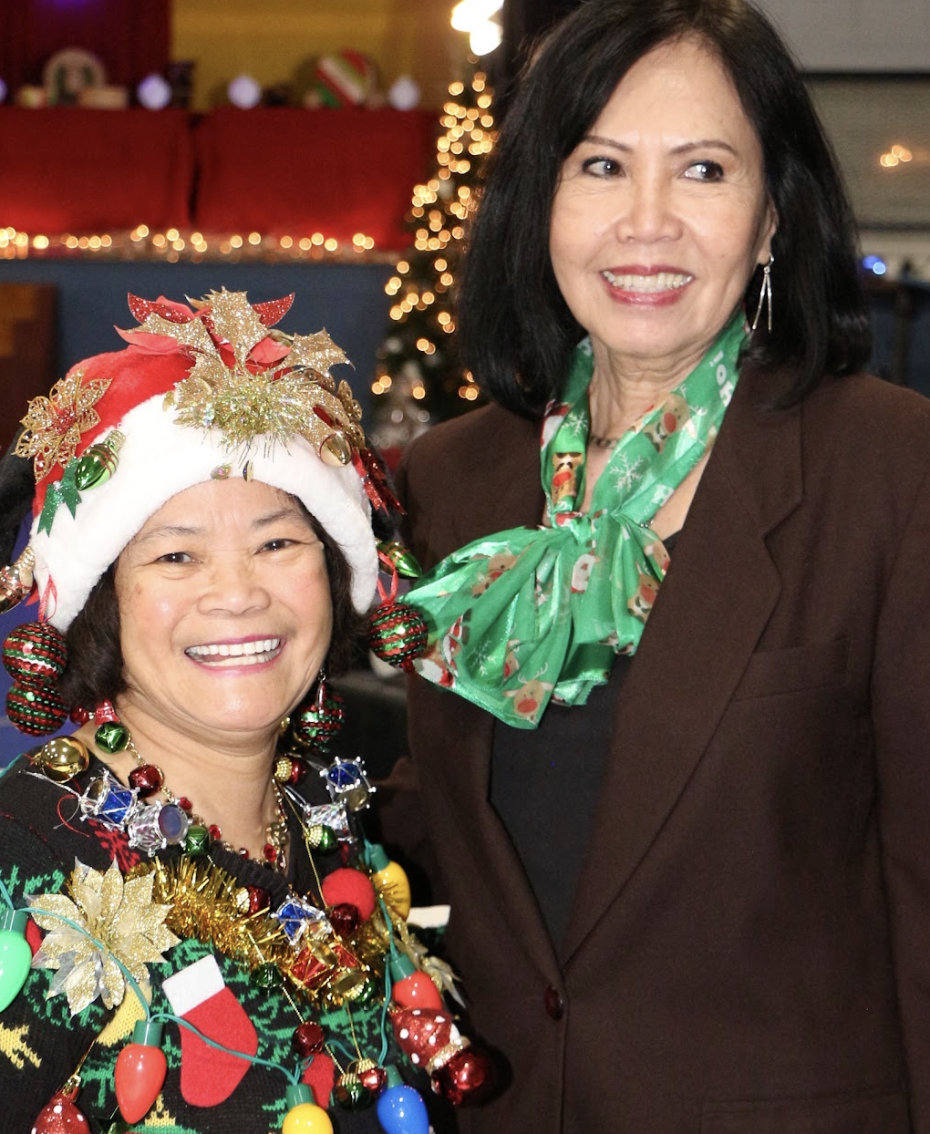 Two women smiling during a Christmas celebration, one wearing a party hat decorated with Christmas ornaments and the other in a brown suit with a green holiday scarf, with Christmas lights in the background.