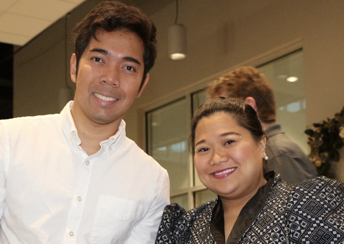 A smiling man in a white button-up shirt standing next to a smiling woman in a patterned black and white jacket, both posing indoors at what appears to be a social event.
