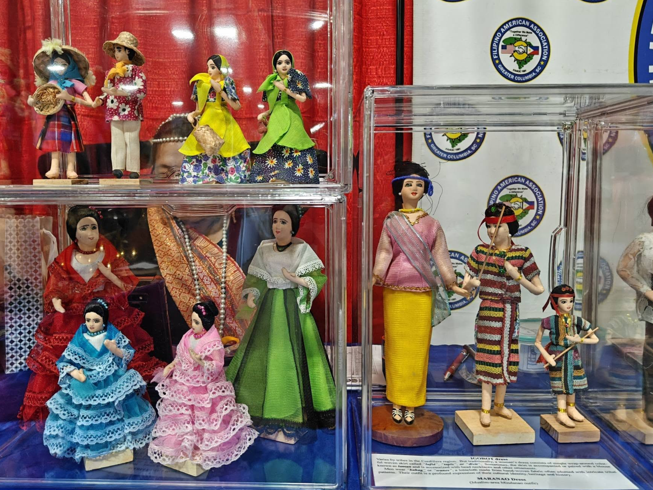 Display of traditional Filipino dolls dressed in colorful clothing on a clear plastic shelf at a cultural exhibit.