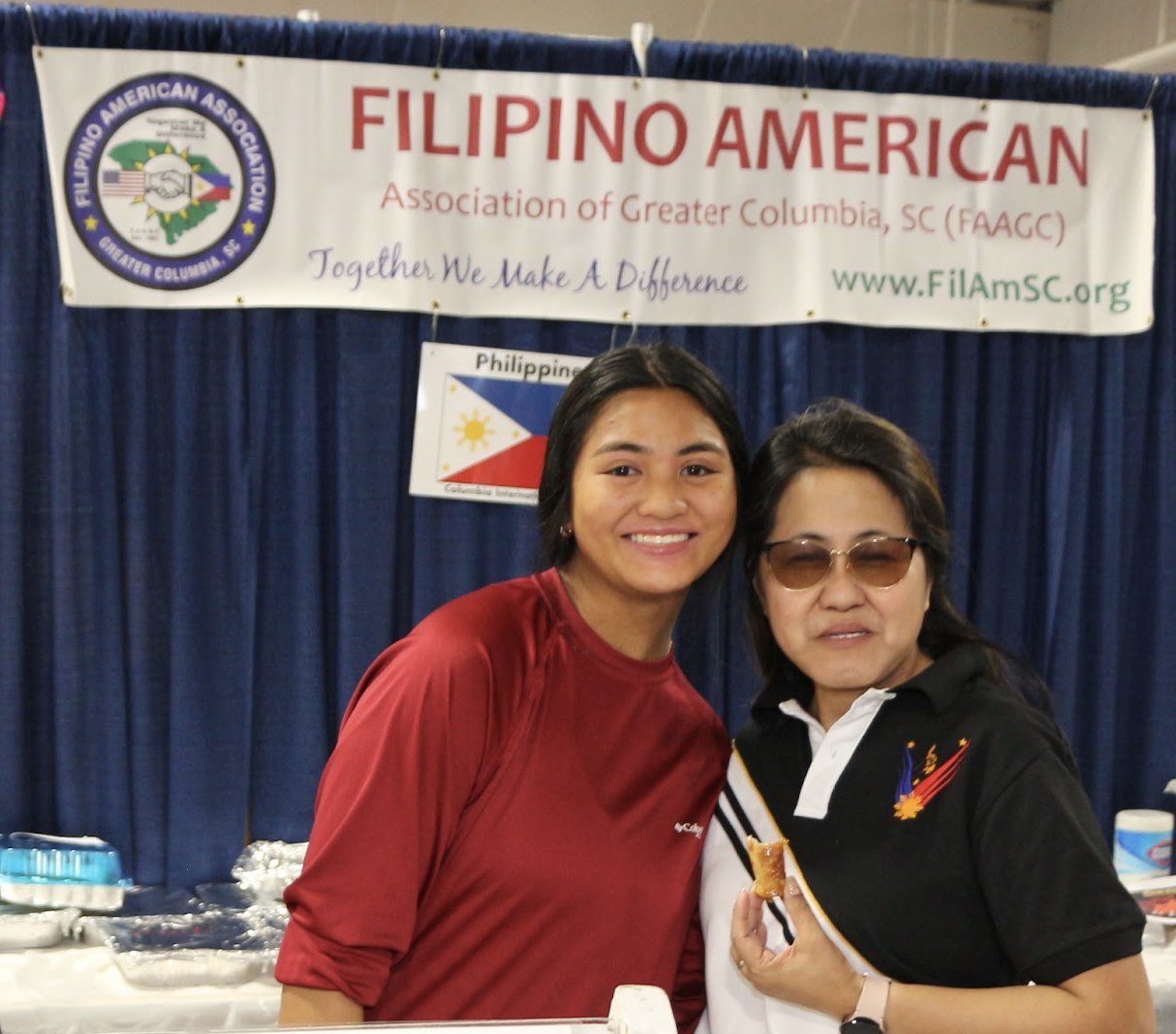 Two women smiling and posing together at a Filipino American association event, standing in front of a banner with the text 'Filipino American Association of Greater Columbia, SC.' The woman on the left wears a red shirt, while the woman on the right