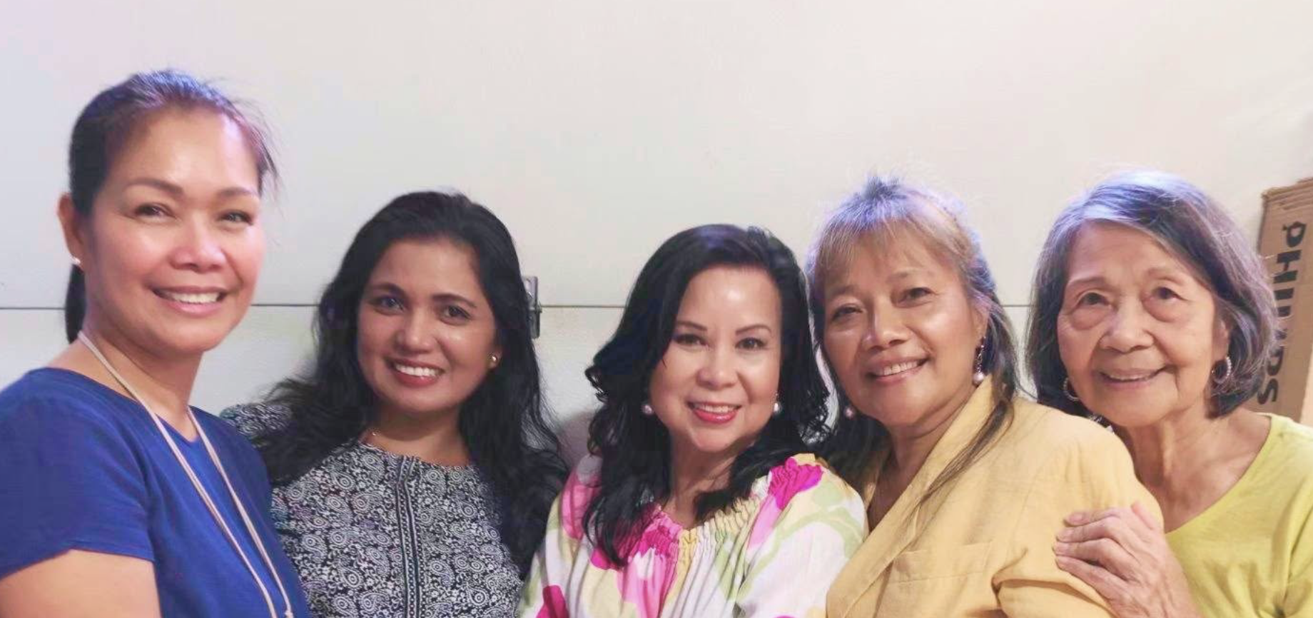 Group of five women smiling indoors, wearing colorful tops, with a plain light background and a cardboard box labeled 'PHIL' partially visible on the right.