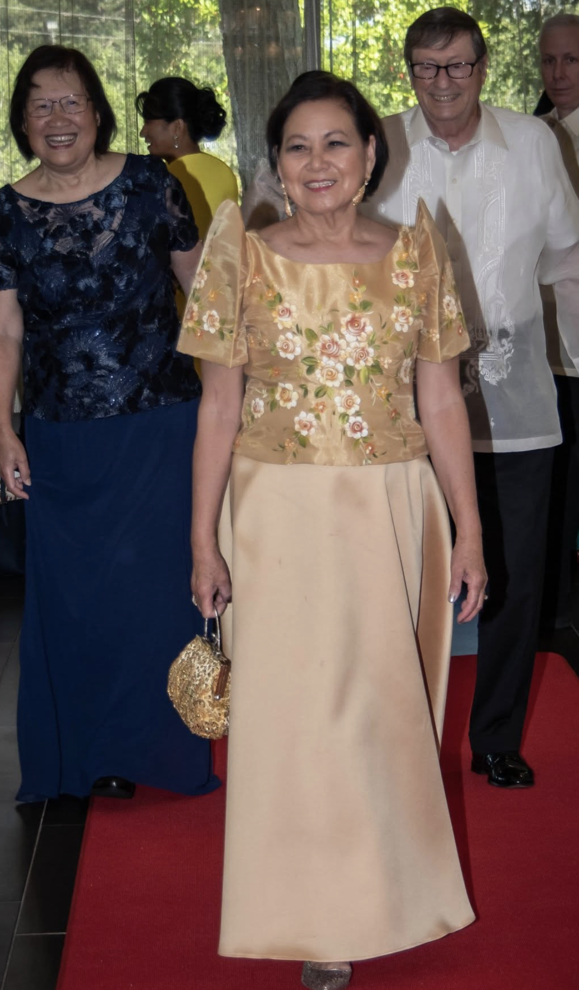 A group of well-dressed people at a formal event, with two women in traditional Filipino attire and two men in formal suits, standing on a red carpet with a green outdoor background visible through a window.