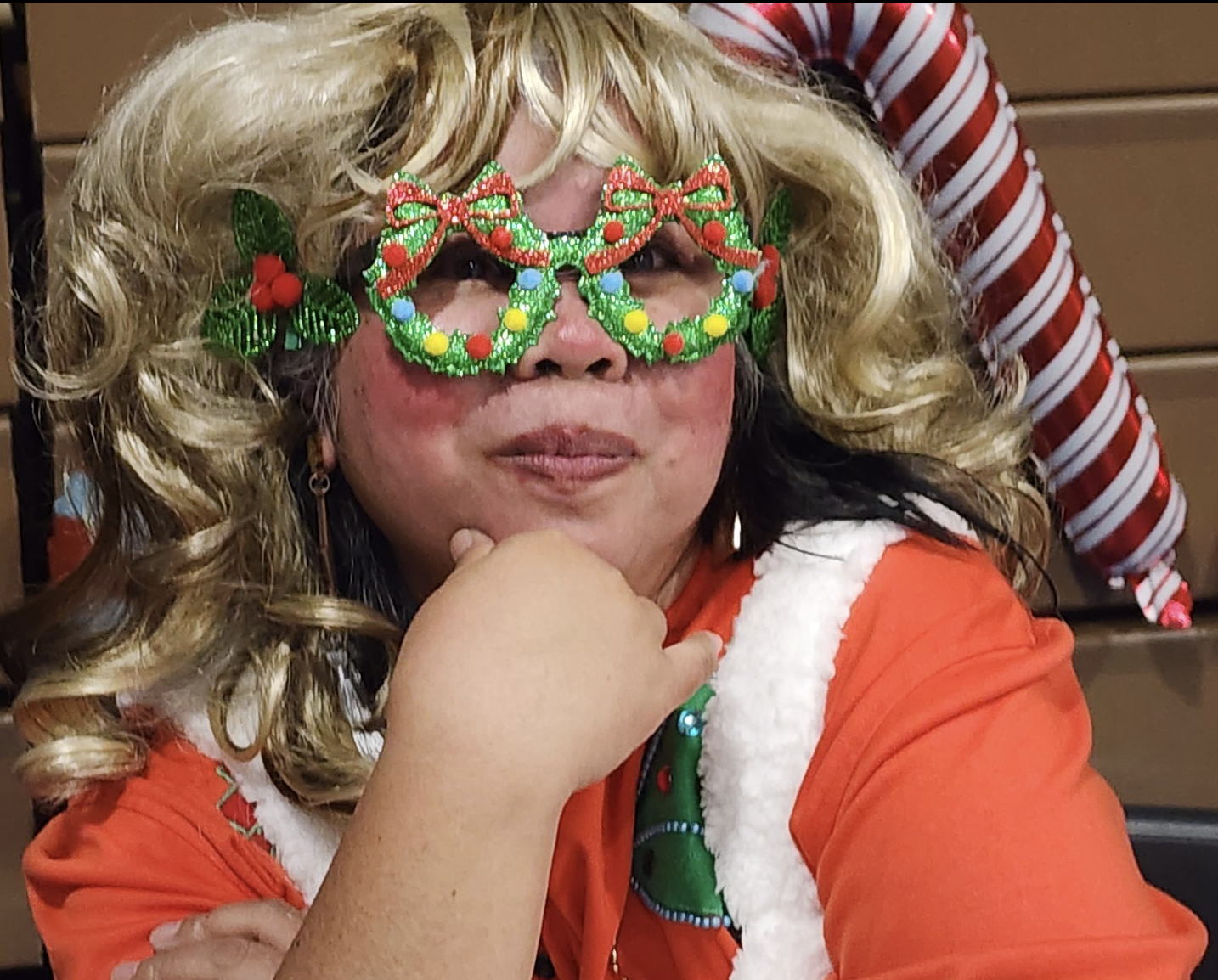 A woman with blonde curly hair, wearing festive Christmas glasses, an orange top, and a Christmas-themed vest, is sitting outdoors with a pained or contemplative expression, resting her chin on her hand.