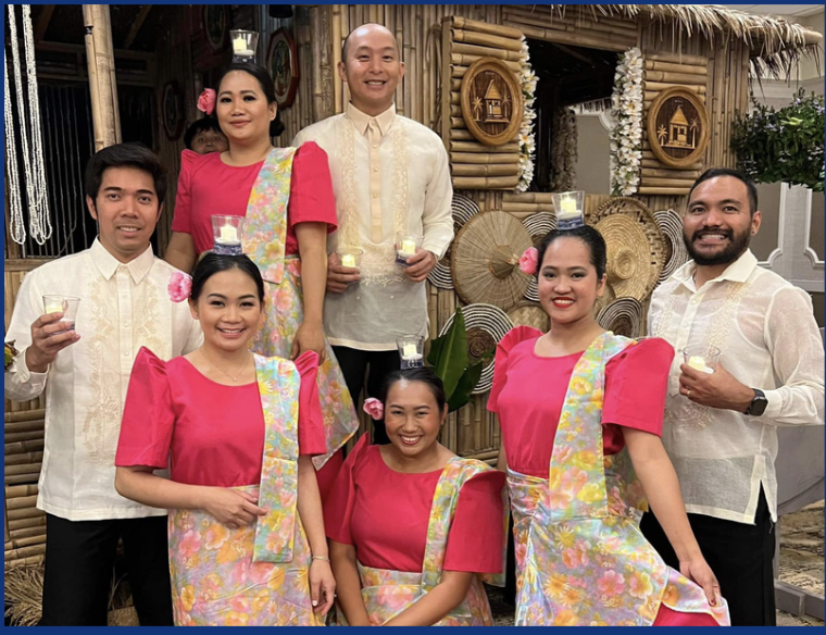 Group of seven people in traditional Filipino attire celebrating, with three women in pink dresses and floral sashes, and four men in cream barongs, holding drinks, in a decorated tropical-themed setting with bamboo and floral decorations.