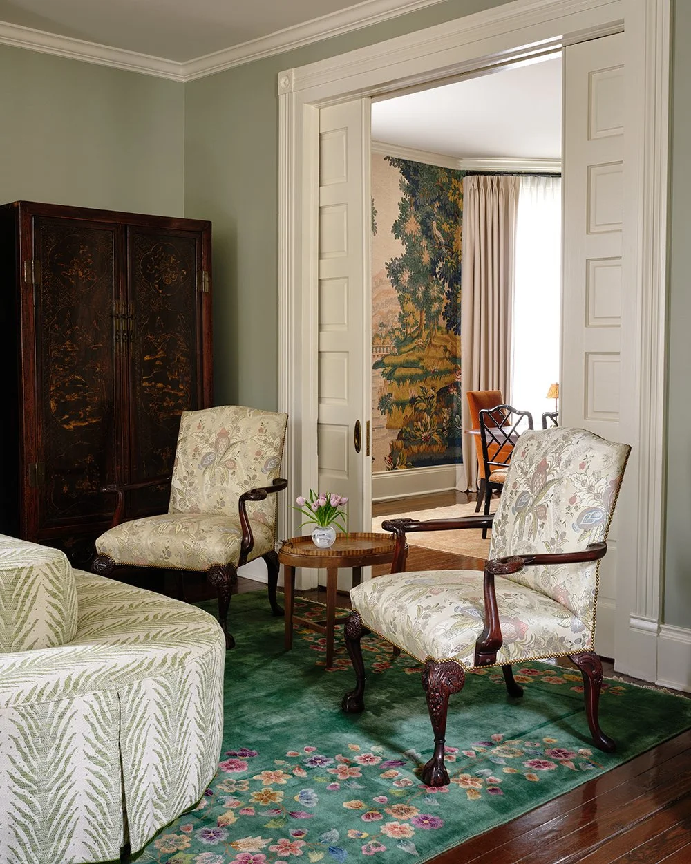 Living room with wooden sliding doors that show a dining room, wooden accent dresser, cushioned antique chairs, and a green floral carpet. 