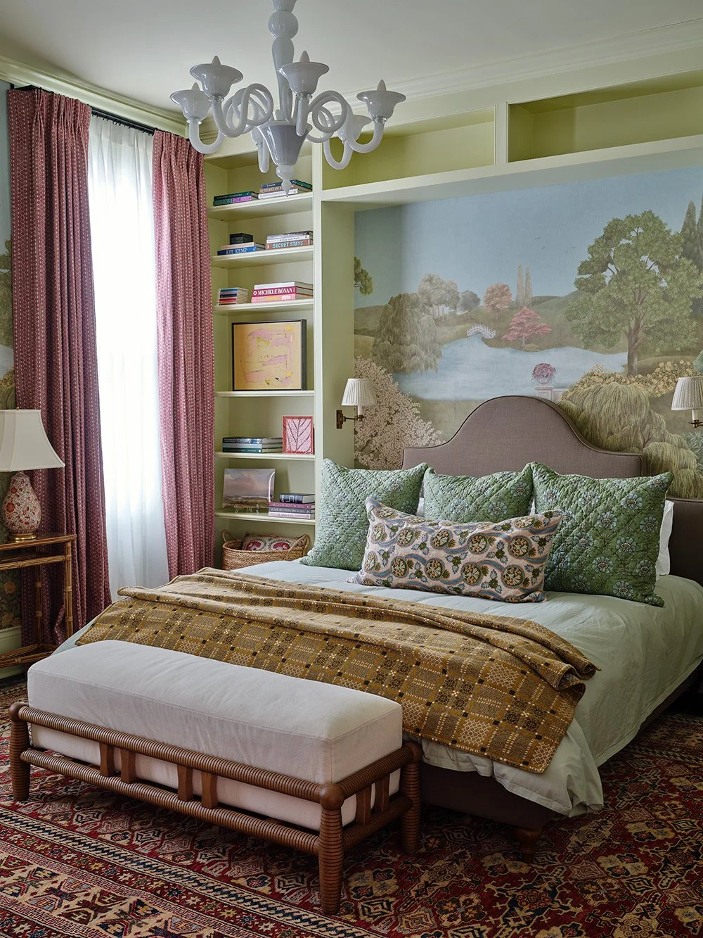 Colorful bedroom featuring green built-in bookshelves, scenic landscape mural, patterned bedding, upholstered headboard, and vintage Persian rug.
