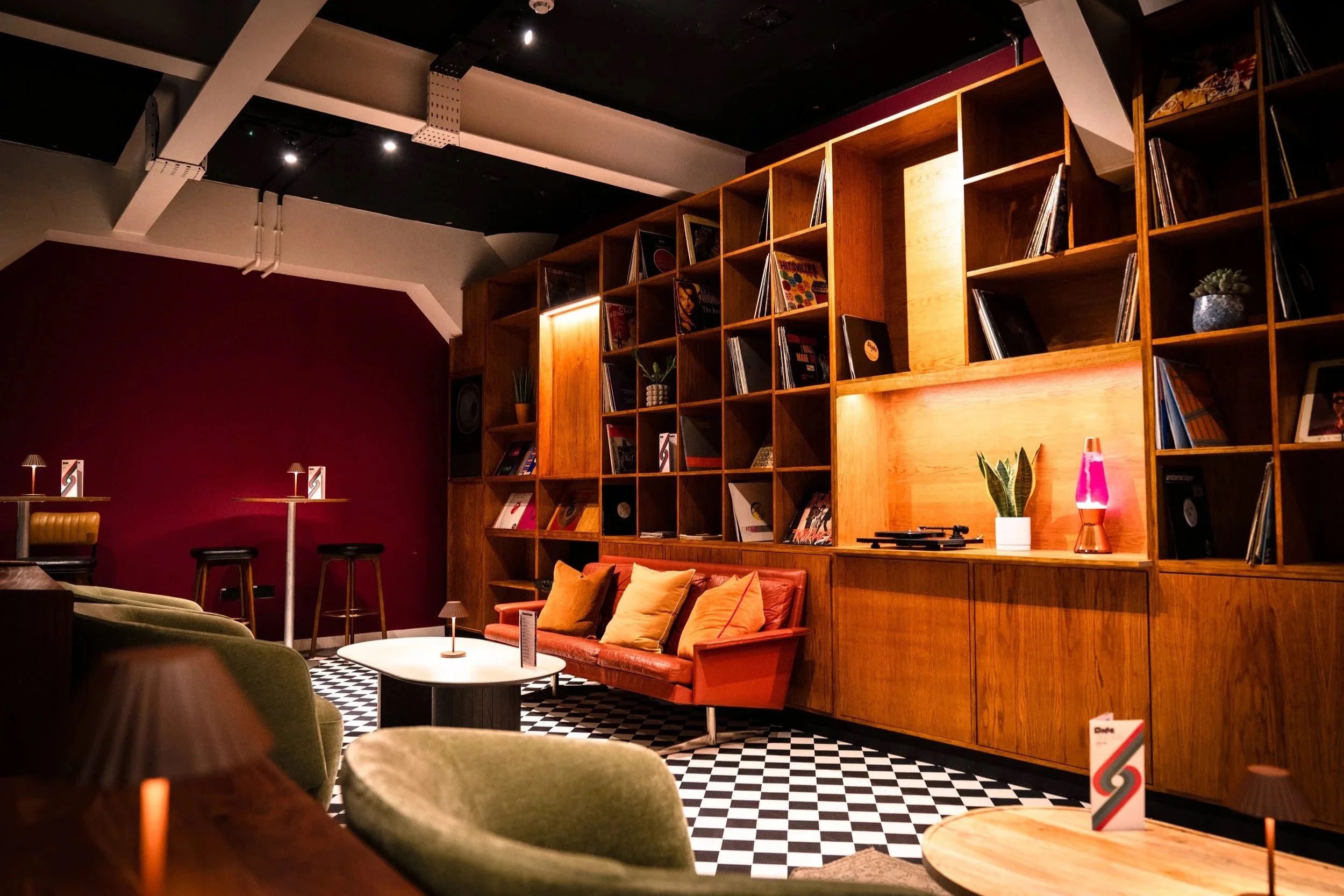 Interior of a cozy lounge with a brick wall, wooden shelves filled with records, a red leather couch with orange and yellow pillows, a low coffee table, green armchairs, and a black-and-white checkered floor.