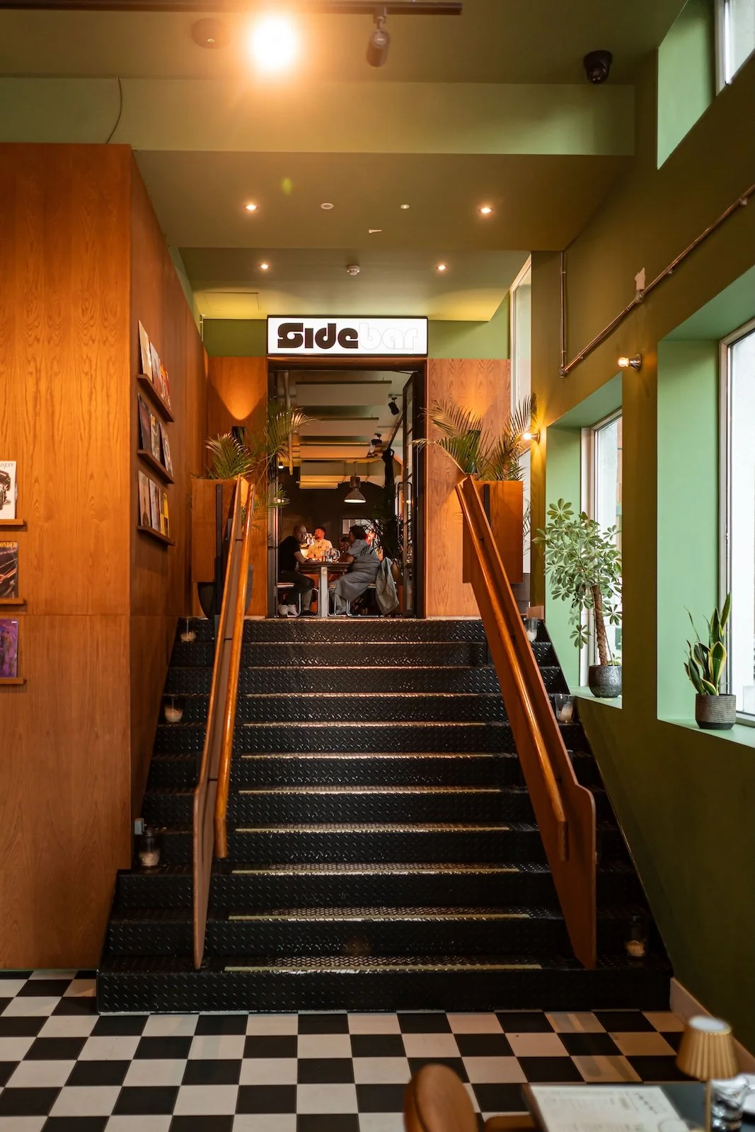 Interior view of a restaurant with stairs leading to a second floor, with people dining inside. The decor includes green walls, wooden accents, and potted plants by the windows.