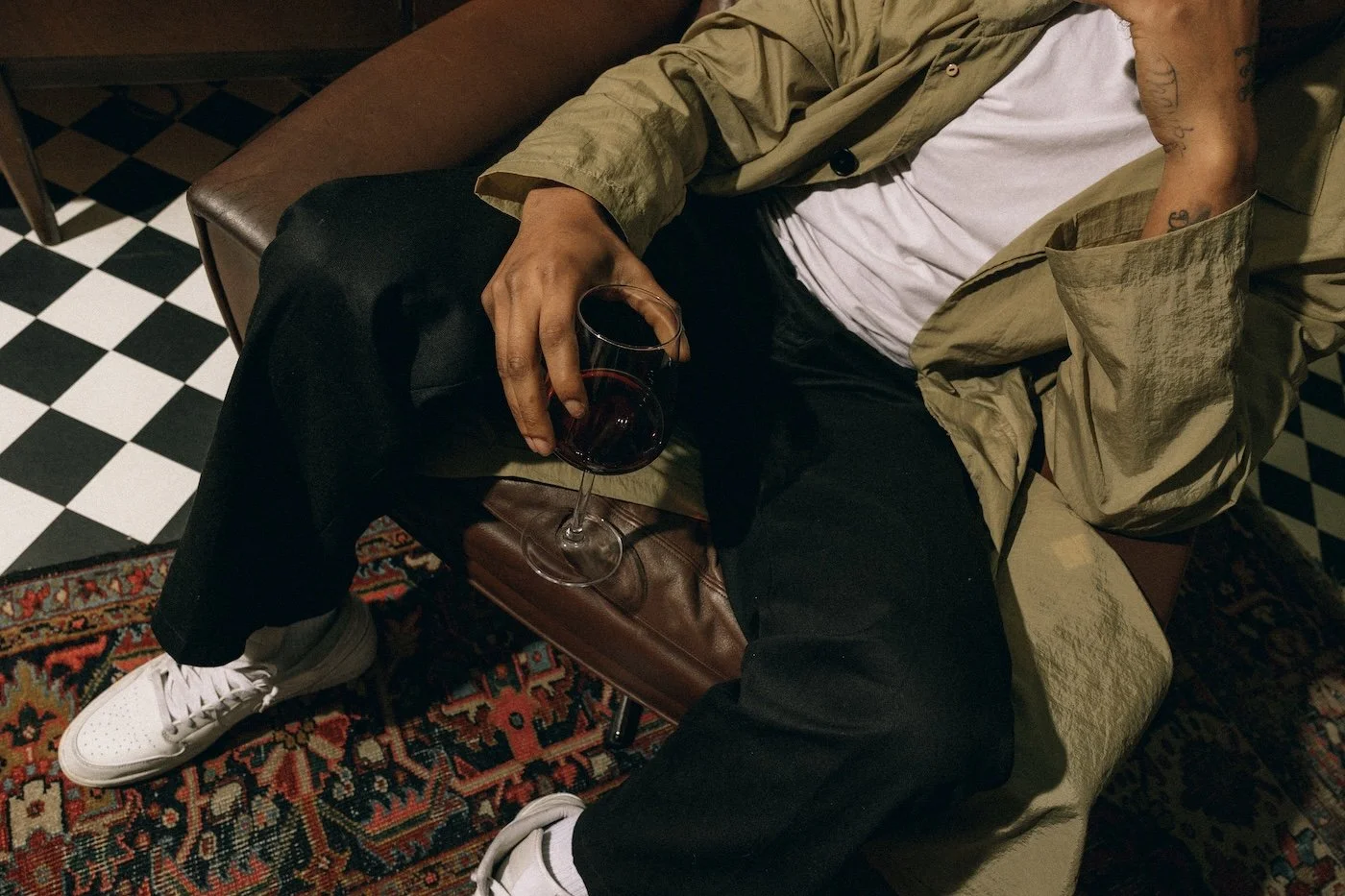 Person sitting on a brown leather couch, holding a glass of red wine in their right hand, wearing a white T-shirt, black pants, white sneakers, a beige jacket, and has tattoos on their left arm. The background features black and white checkered flooring and a patterned rug.