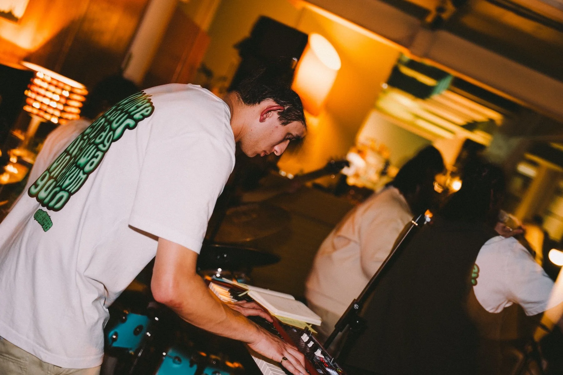 A young man with dark hair playing a keyboard or synthesizer at a dimly lit indoor event or party, with warm yellow lighting and blurred background of other people.