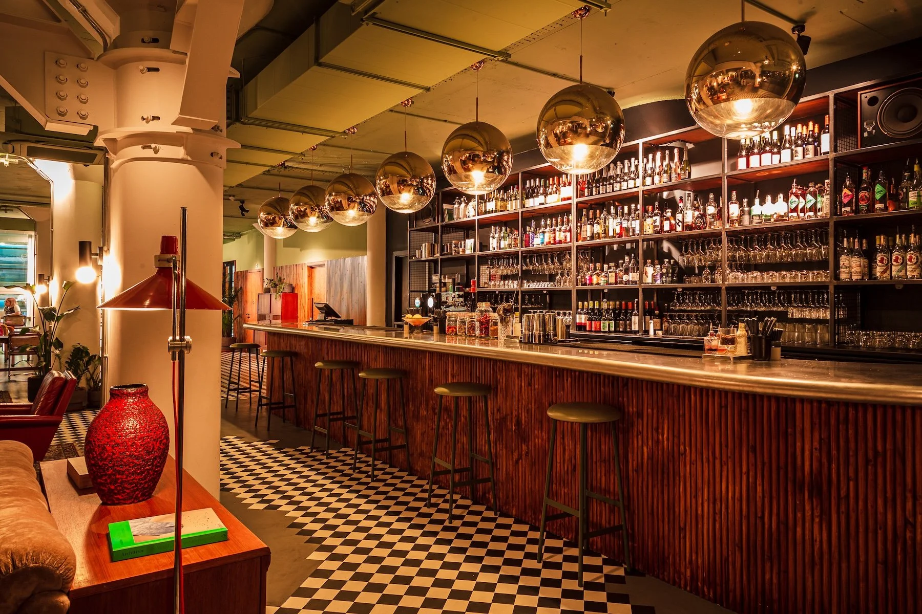 Inside a modern bar with a long wooden counter, shelves stocked with liquor bottles, large gold hanging spheres, checkered black and white floor, and seating stools.