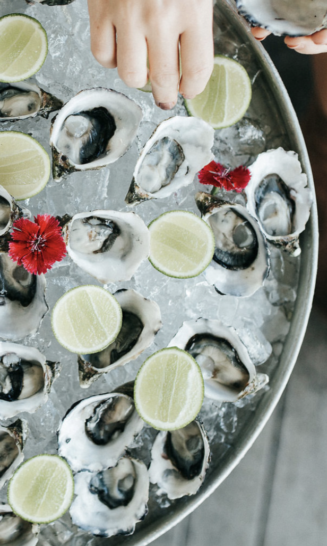 A tray of fresh oysters on ice garnished with lime wedges and red flowers, with a hand reaching for an oyster.