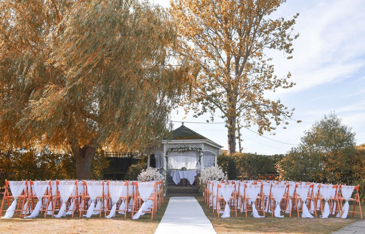 Outdoor wedding setup with rows of white chairs decorated with white sashes, leading to a gazebo adorned with white flowers and drapery, under tall trees with autumn leaves.