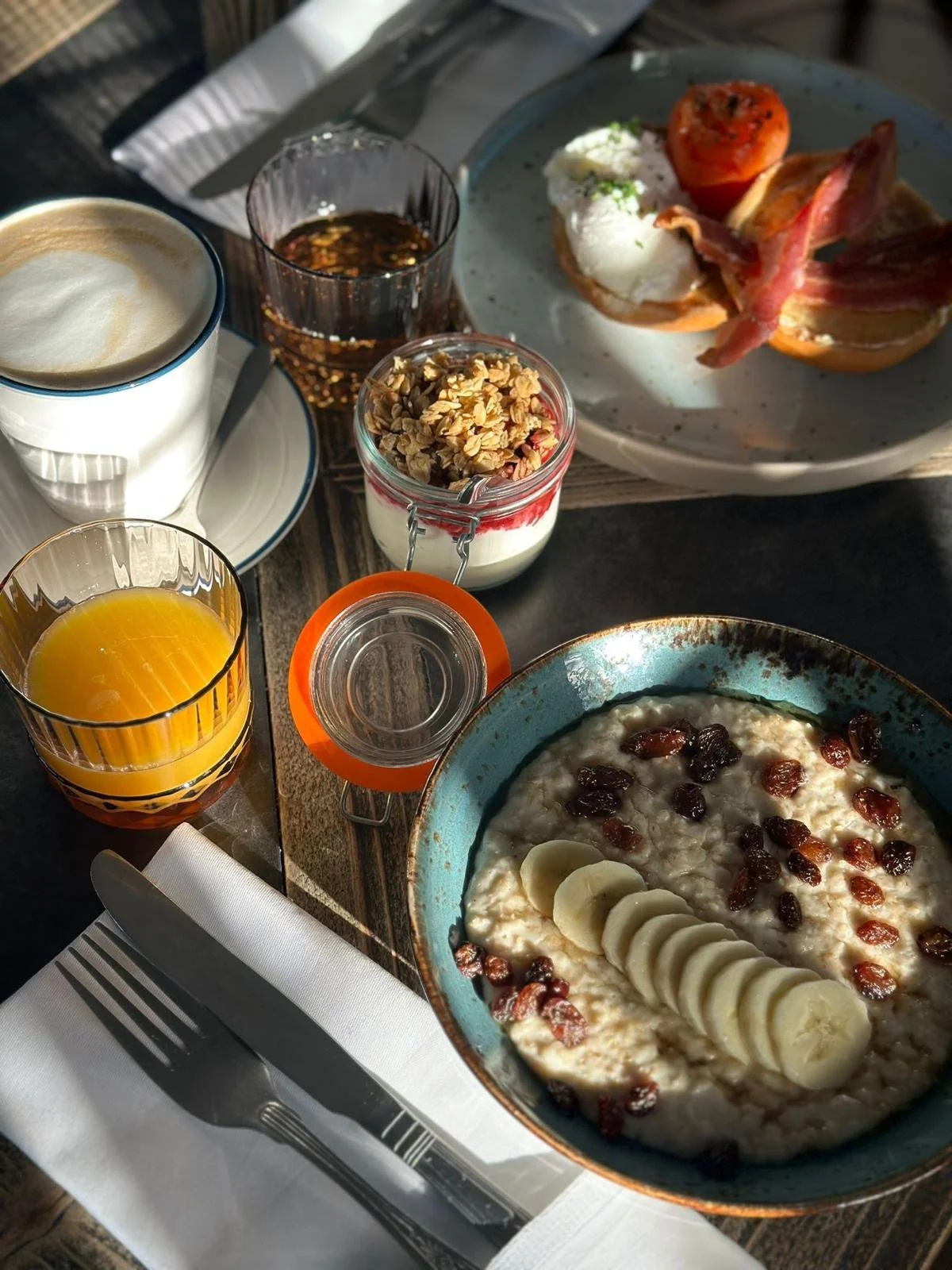 Breakfast spread with oatmeal topped with banana slices and raisins, orange juice, coffee, yogurt with granola and berry compote, a glass of syrup, coffee, and a plate with tomato, poached egg, bacon, and toast on a wooden table.
