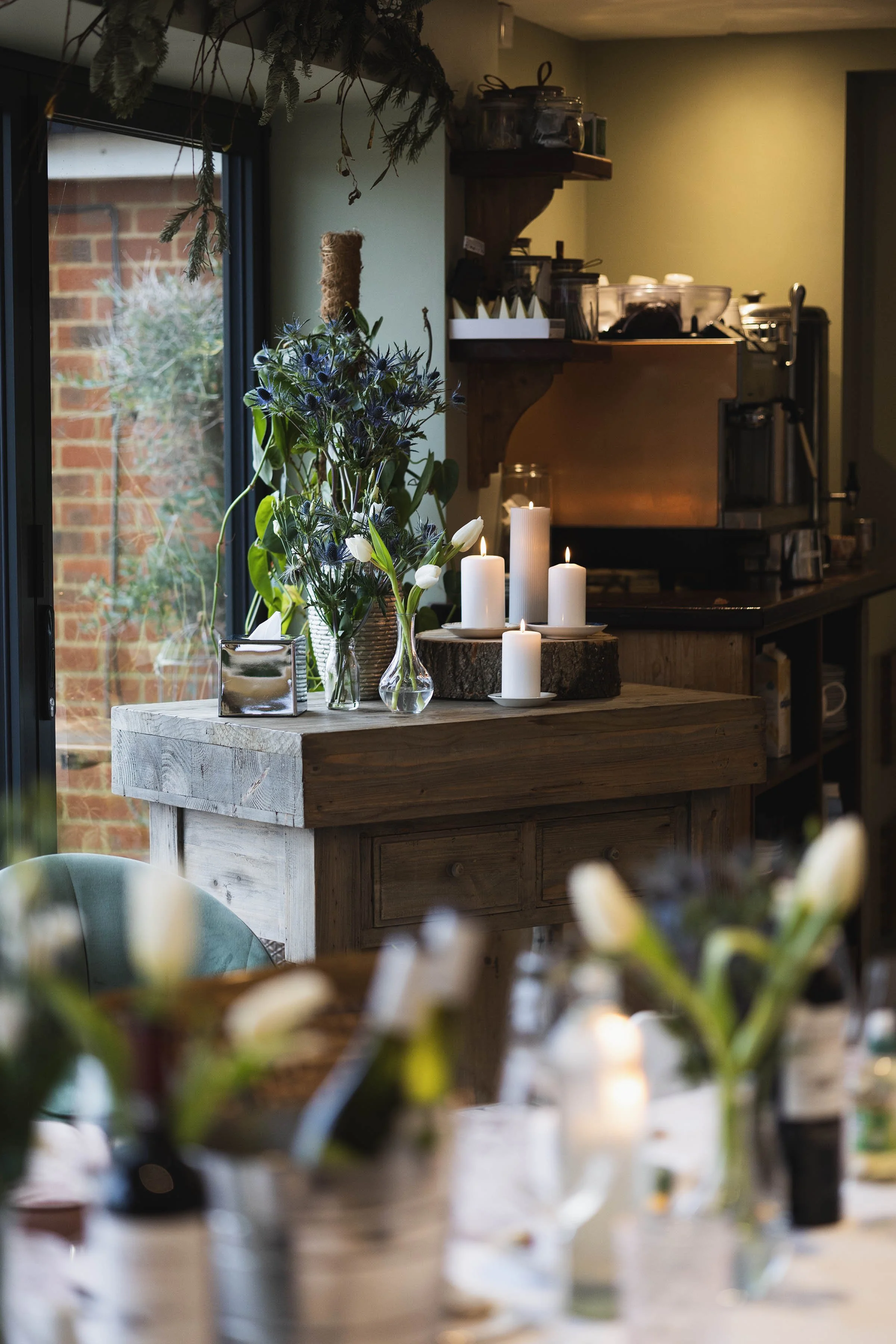 A cozy kitchen or dining room with a wooden sideboard, vases of flowers, and lit candles, near a sliding glass door that reveals a brick exterior.