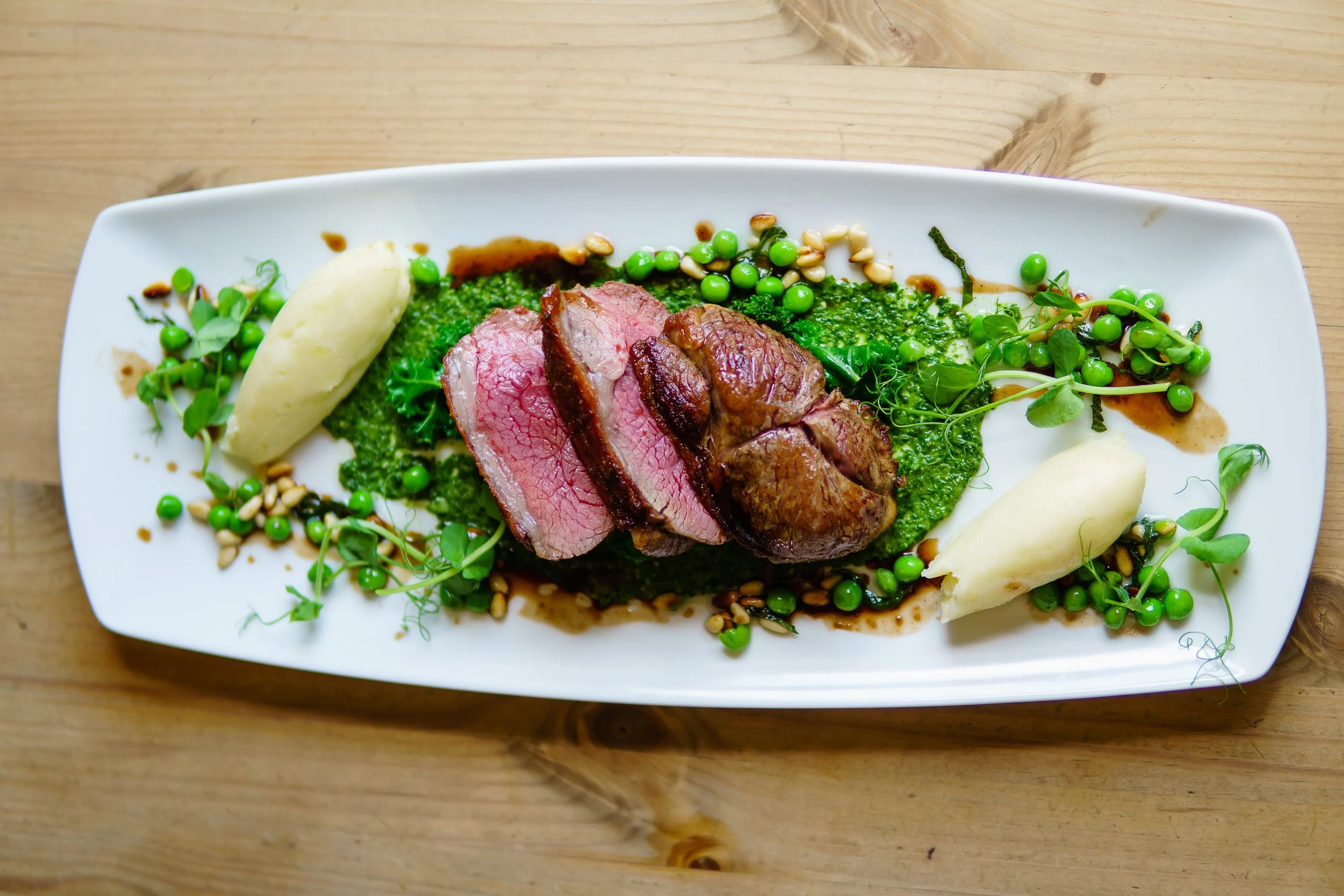 Plate of sliced cooked beef, mashed potatoes, green peas, leafy greens, and microgreens, with sauce on a white rectangular plate.