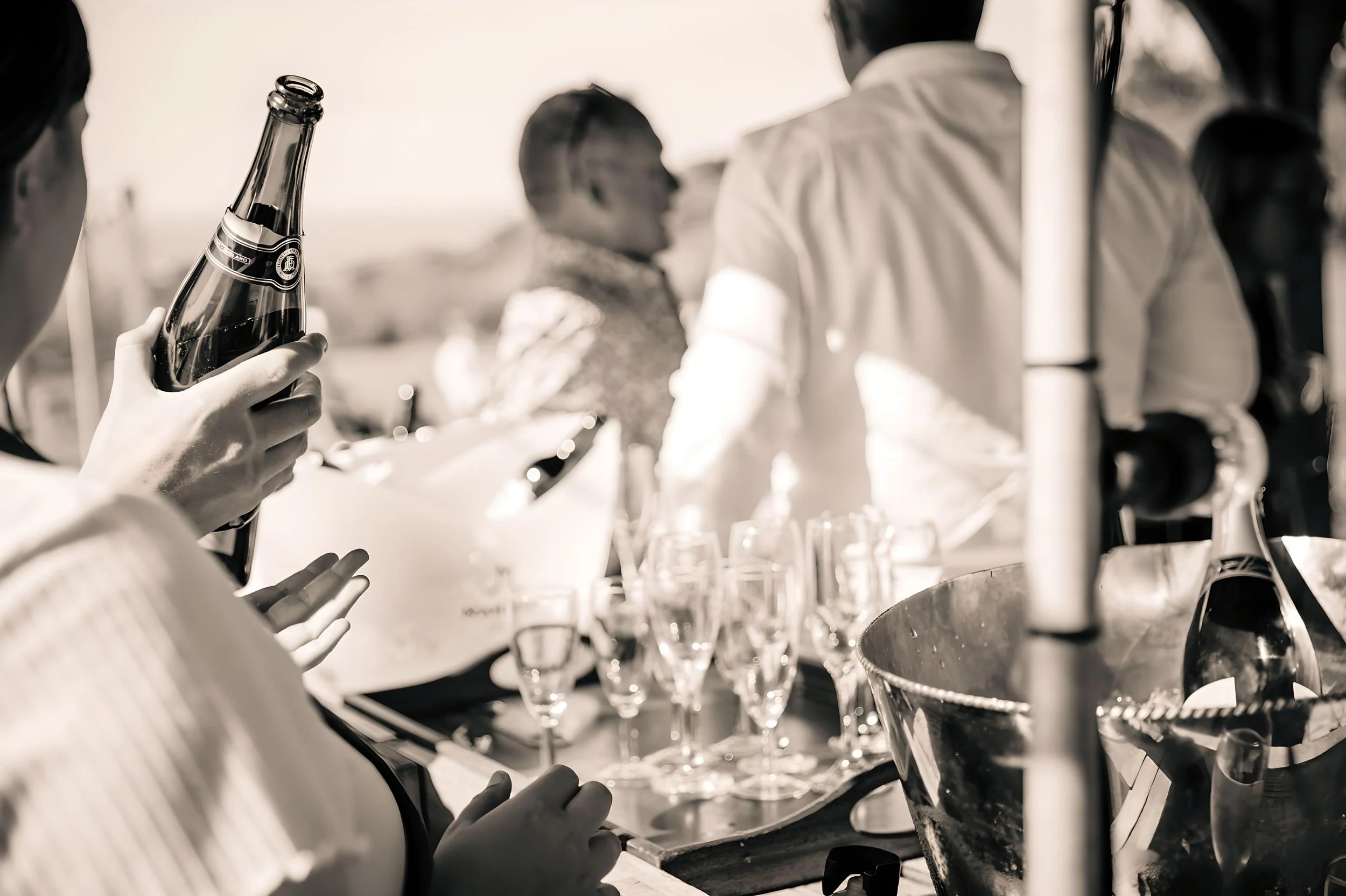 A person holding a glass bottle of beer with others nearby. Several glasses and a bottle are on a table, with people socializing in the background, outdoors, in black and white.