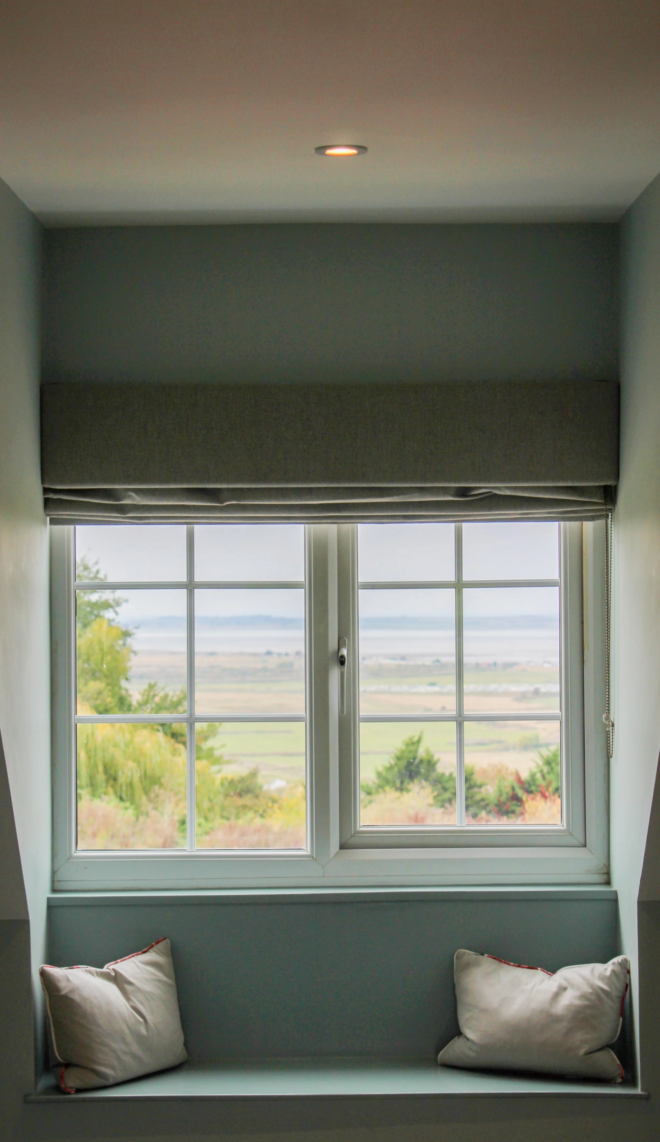 A window seat with two white pillows below a large window with a view of trees and water in the distance.