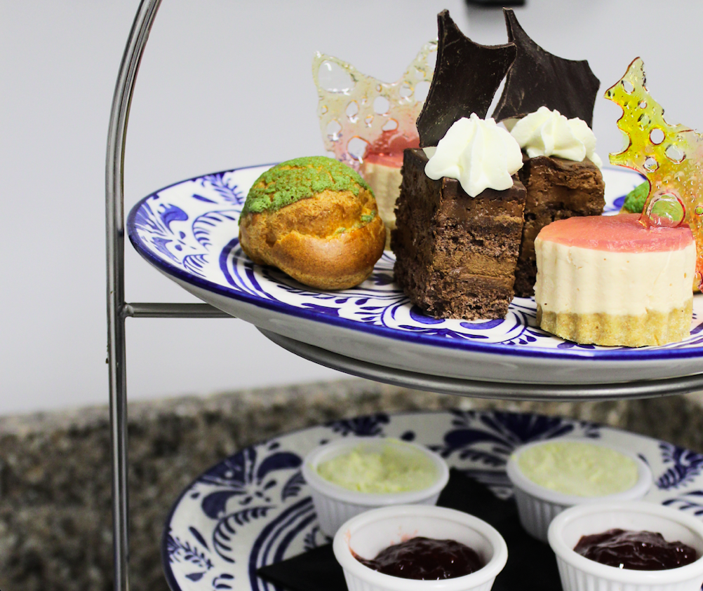 A two-tiered dessert stand holding various desserts including a green-topped muffin, a chocolate cake with white frosting, a pink-topped cheesecake, and decorative sugar pieces, with small bowls of cream and jam underneath.