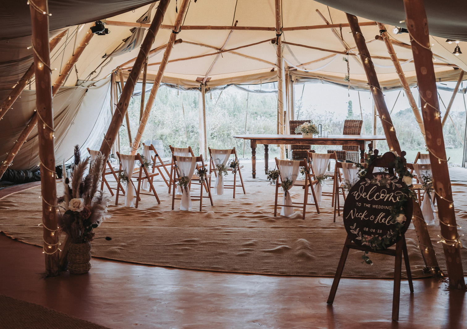 Interior of a rustic wedding tent with a circular arrangement of chairs decorated with white fabric and small flower bouquets, a welcome sign on an easel, a table in the background with floral arrangements, and natural greenery outside the tent.
