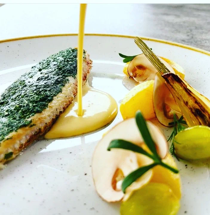 A plated dish featuring a piece of fish with crispy skin, topped with a yellow sauce, alongside roasted pineapple slices, green grapes, and garnished with herbs and edible flowers.