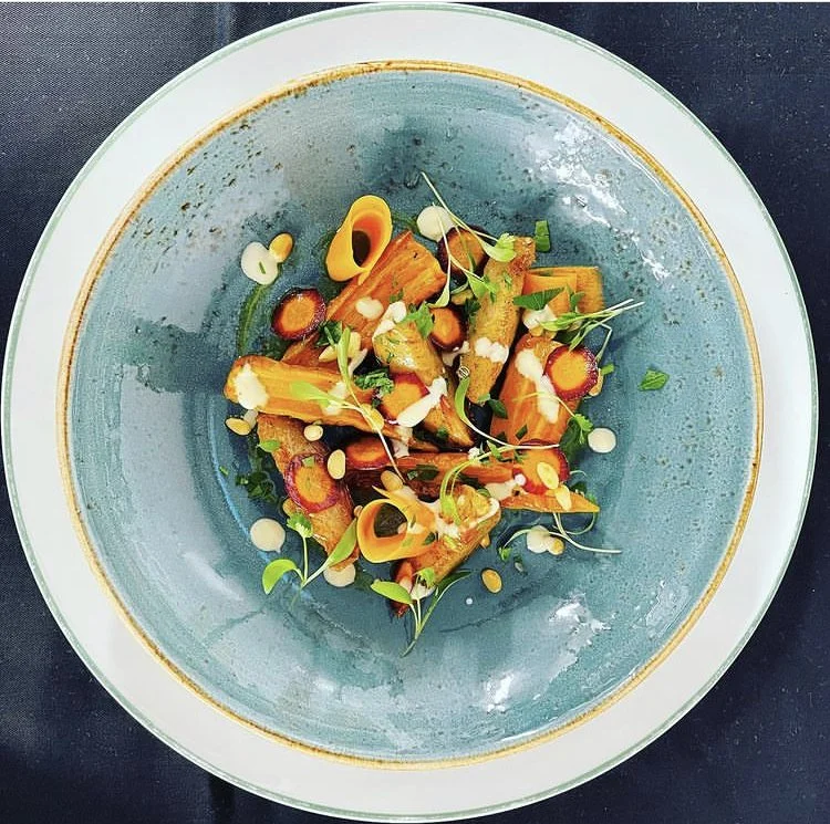 A plated gourmet dish with sliced vegetables, microgreens, and sauce drizzles on a blue ceramic plate.