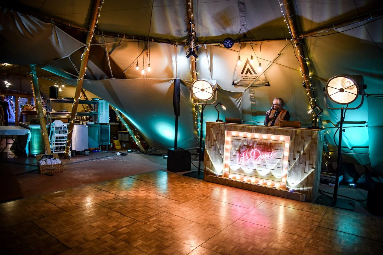 Interior of a decorated event tent with a wooden dance floor, a DJ booth with a sign that says 't Dance', decorative lighting, and a DJ man wearing glasses.