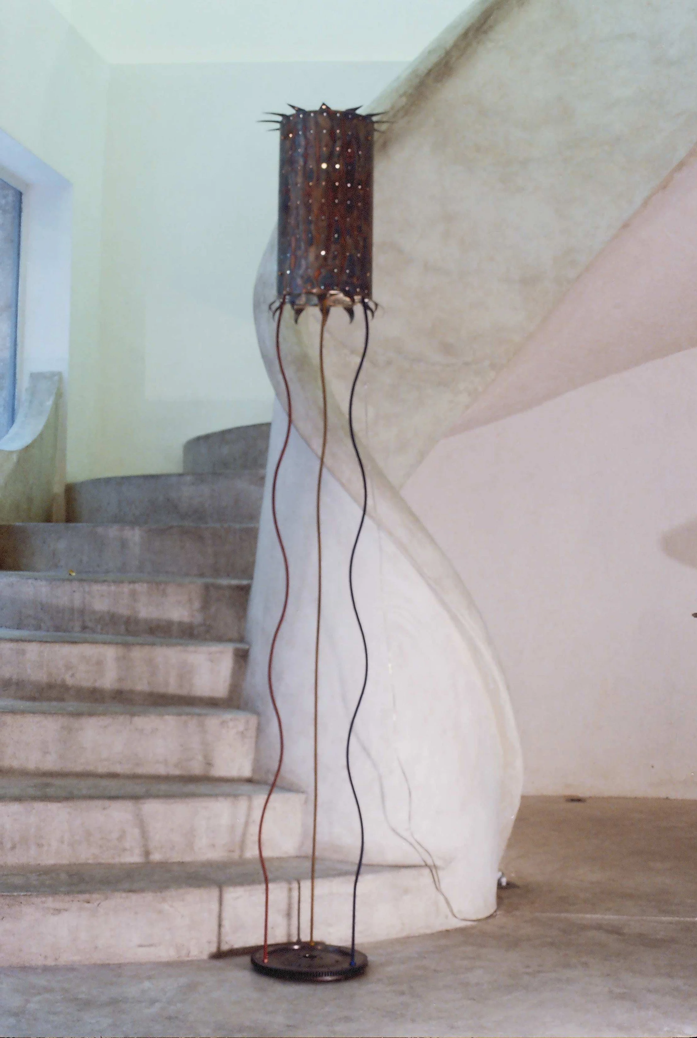 A decorative lamp with a dark, spiked shade and protruding wires, standing beside a staircase with a rounded, textured wall.


