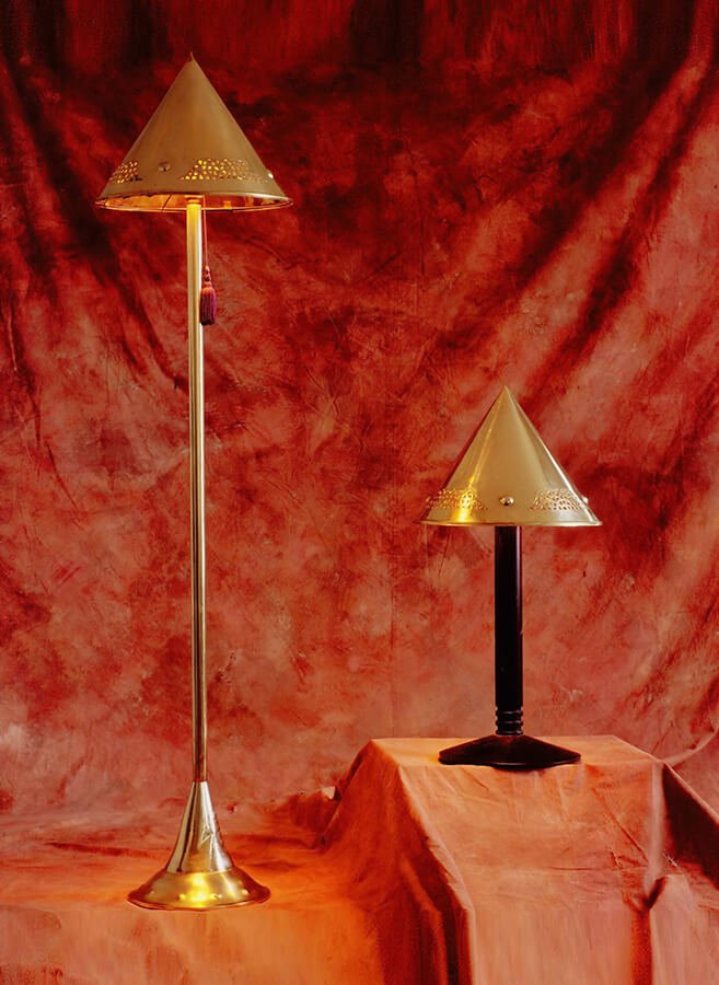 Two decorative table lamps with gold cone-shaped metal shades, one tall and one short, are placed on an orange draped surface against a red textured backdrop.


