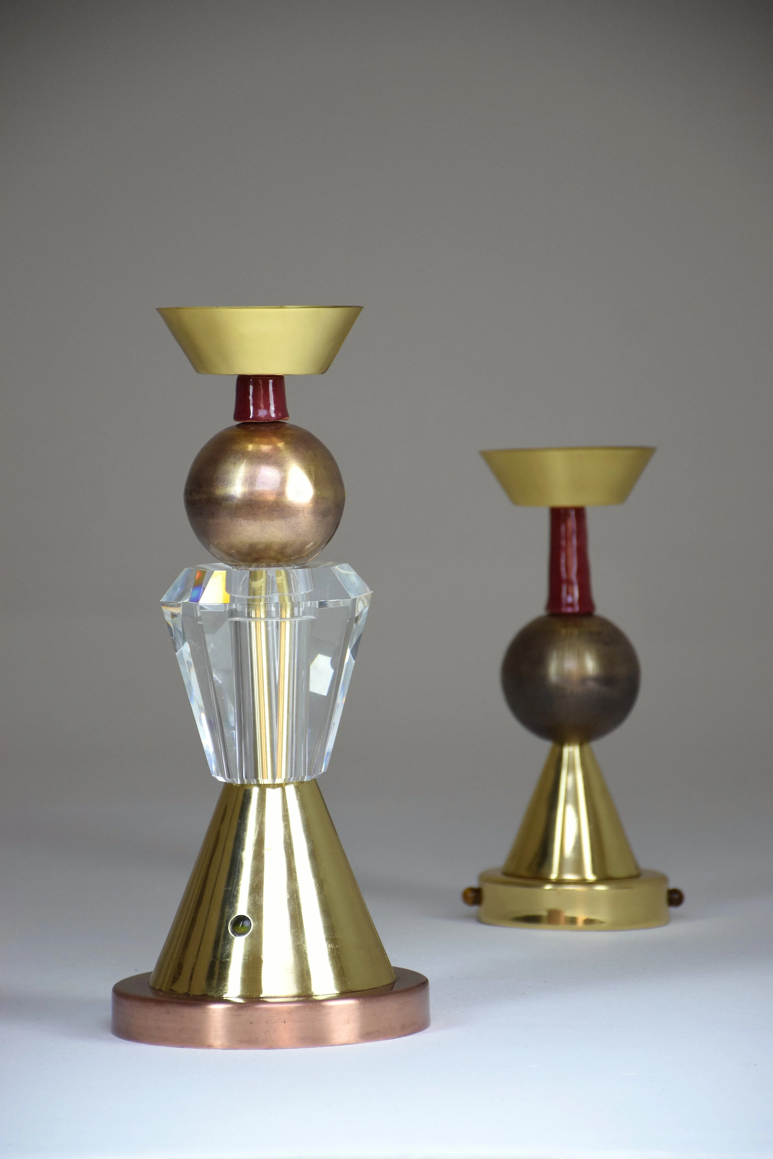 Two modern candlesticks with metallic and glass elements, one in the foreground and one in the background, on a white surface.


