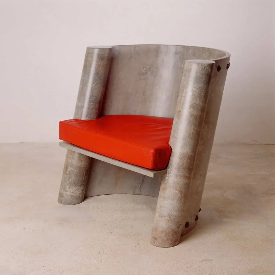 A modern armchair with a concrete-like finish, featuring large cylindrical armrests and a red cushion on the seat, set against a plain white background.


