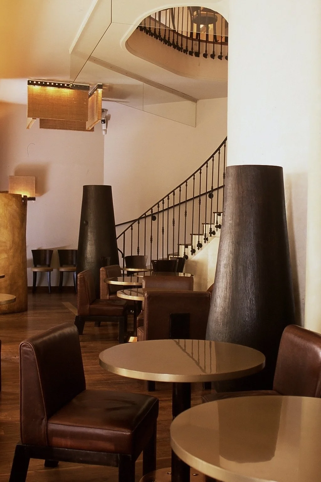 Interior of a restaurant or cafe with round tables, leather chairs, large black cone-shaped decor pieces, a wooden staircase with a metal railing, and warm lighting fixtures.