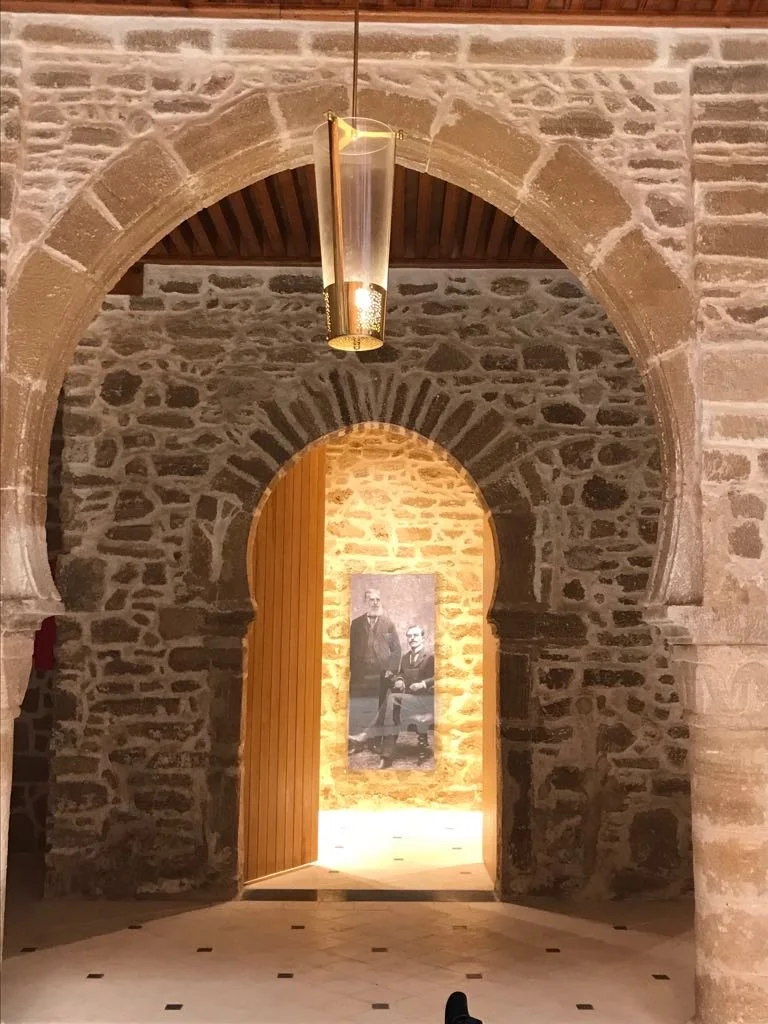 Stone archway leading into a room with a wooden door and a black-and-white photograph of two people, one sitting and one standing, on the wall illuminated with warm lighting.


