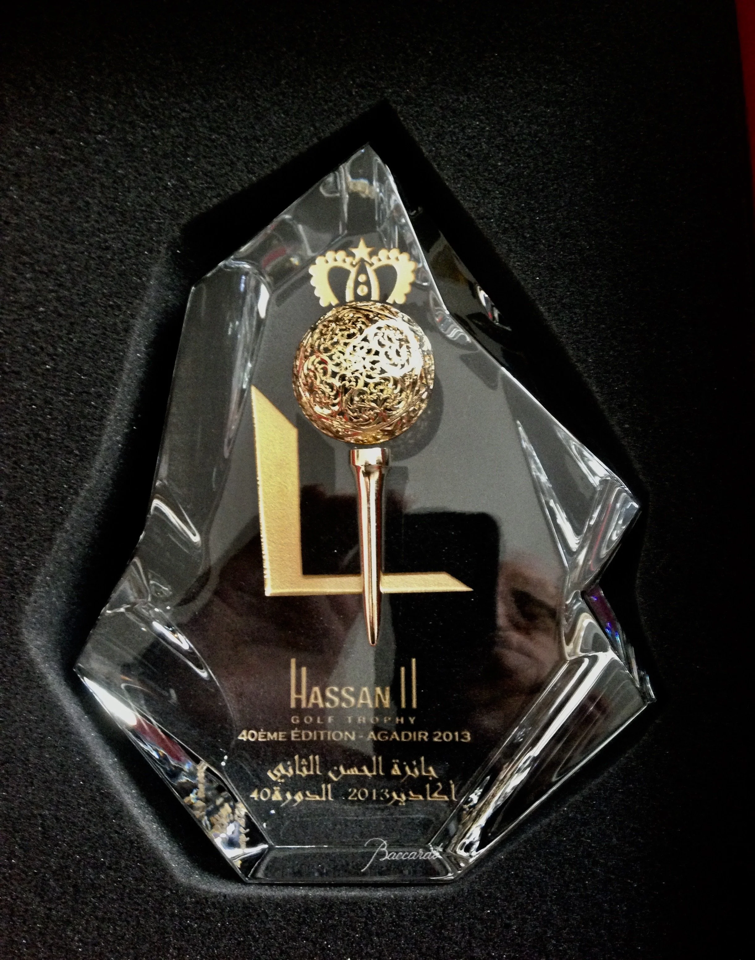 A crystal trophy with a gold pin-shaped decoration and a gold crown symbol on top, engraved with 'Hassan II Golf Trophy 40th Edition - Agadir 2013' in French and Arabic, resting on black velvet.


