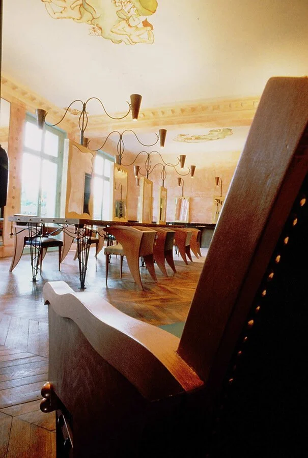 Empty dining room with wooden chairs and tables, large windows, and decorative ceiling artwork.


