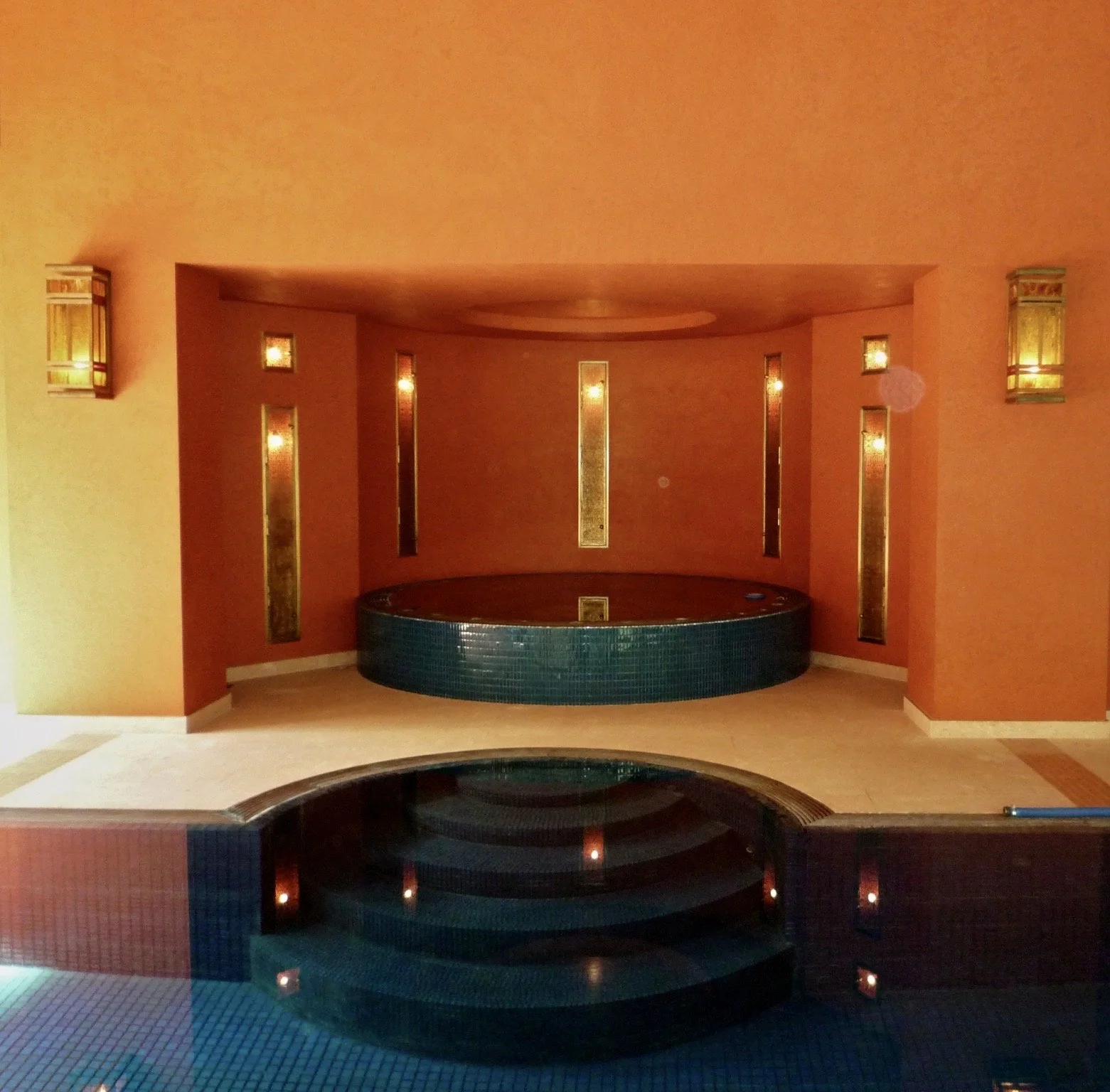 Indoor spa area with a small raised hot tub in front, surrounded by walls with vertical decorative windows and wall sconces with warm lighting.


