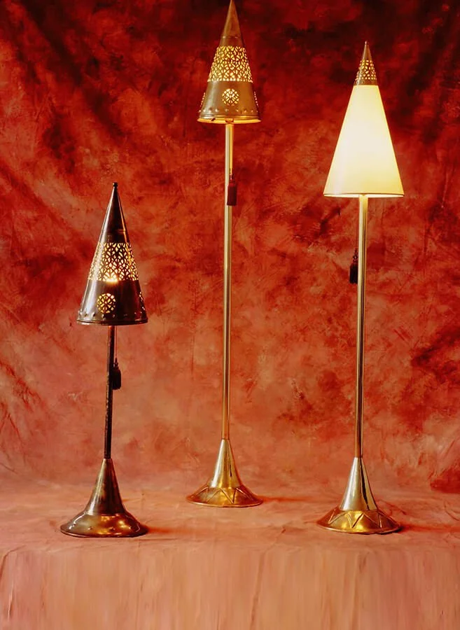Three decorative floor lamps with metallic bases and cone-shaped shades, one black with perforated patterns, one gold with perforations, and one white, all standing on a peach-colored surface against a textured red and orange background.



