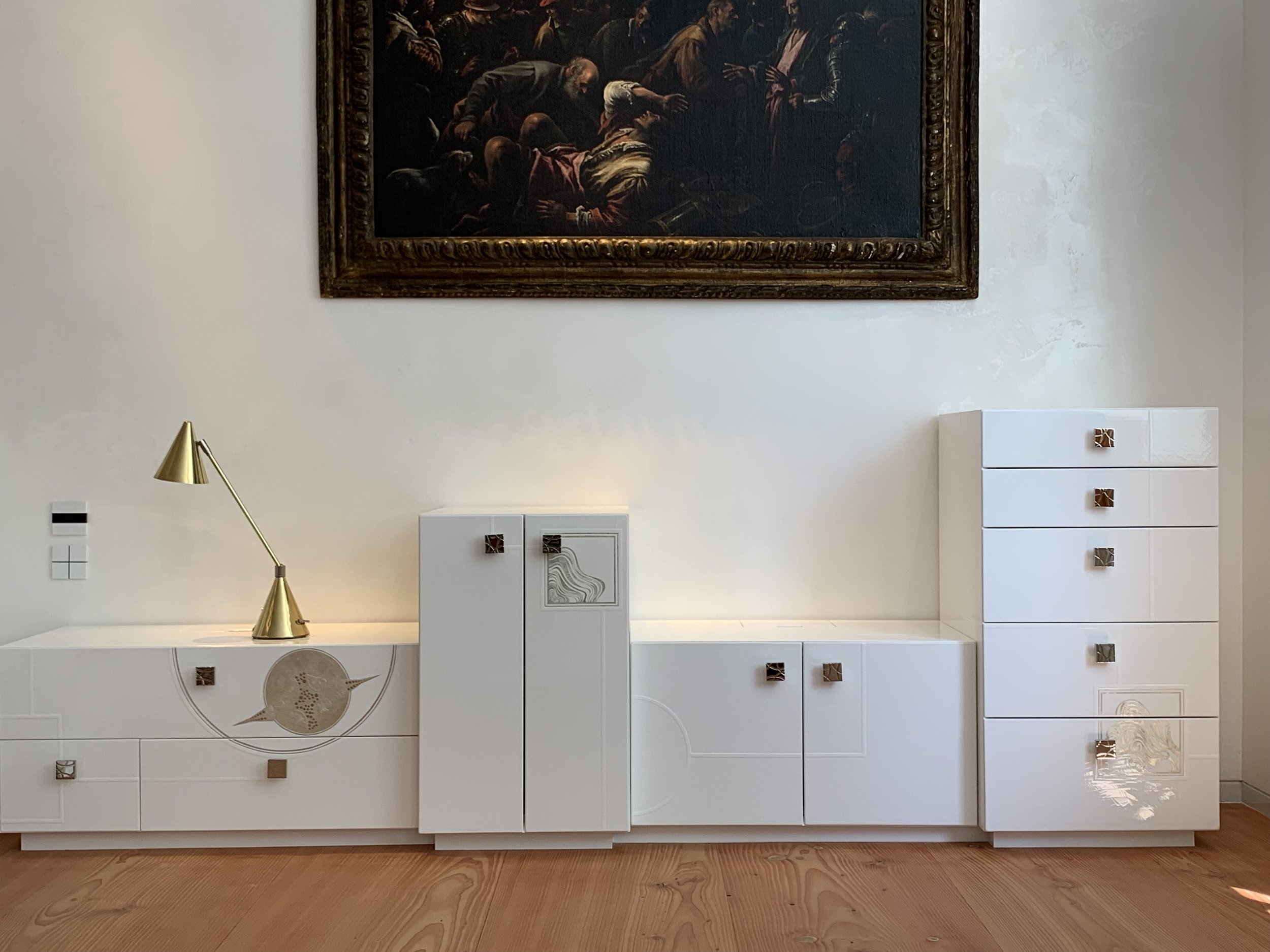 A modern white cabinet with decorative handles and a gold desk lamp on top, set against a light wall with a framed classical painting hanging above.


