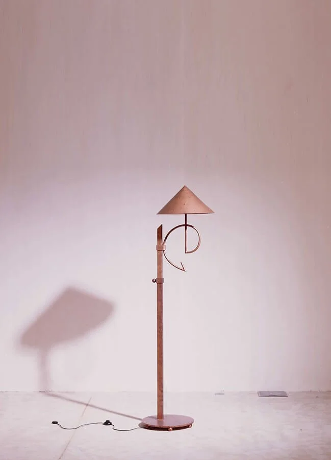 Minimalist floor lamp with an adjustable arm and conical lampshade, casting a shadow on a plain white wall.


