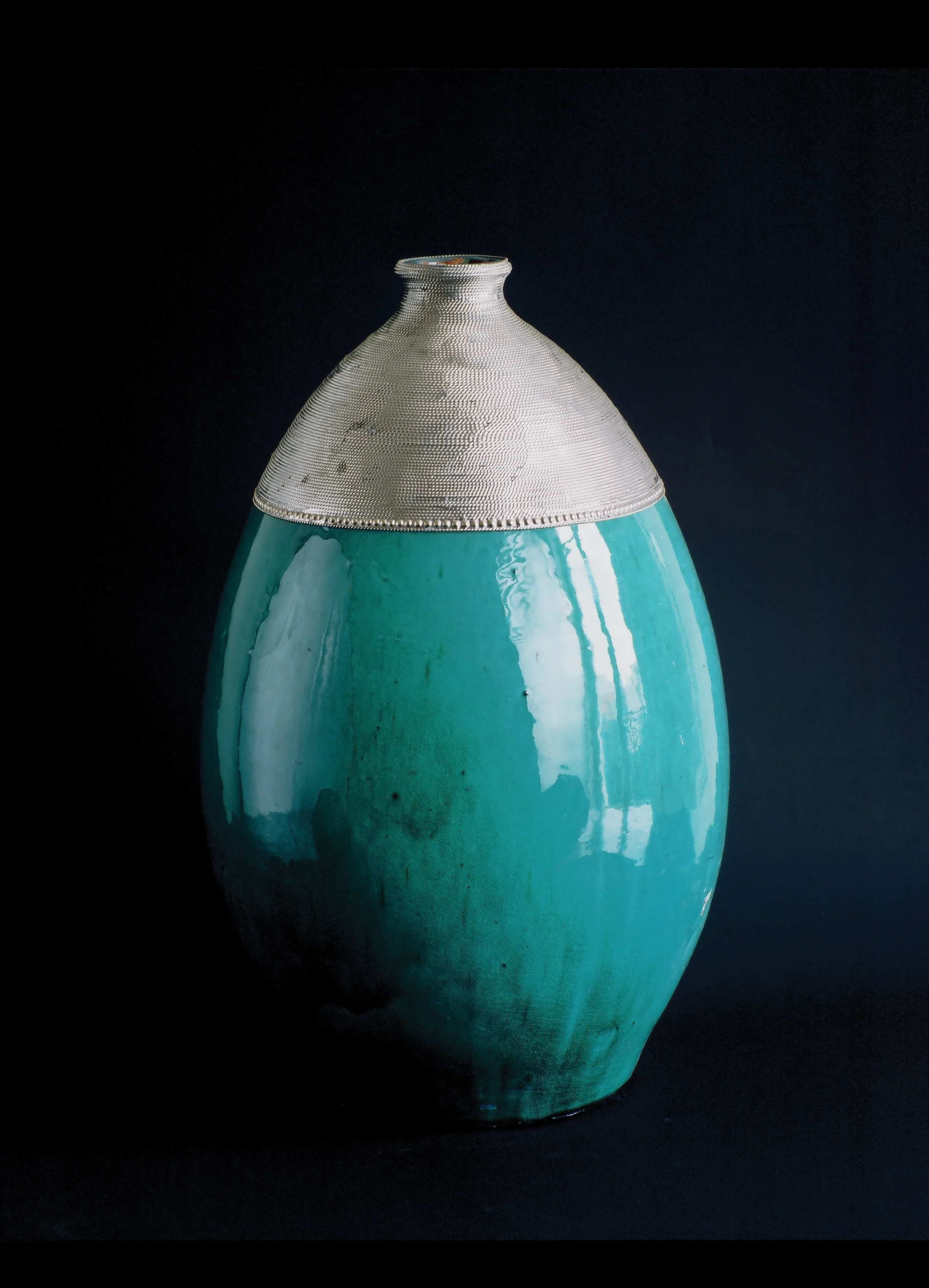 Ceramic vase with a turquoise lower half and a silver metallic upper half, set against a black background.


