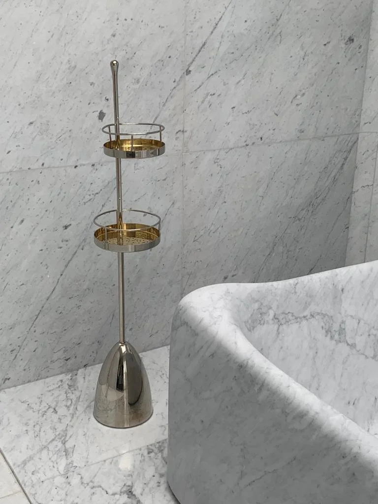 A silver and gold two-tier shower caddy with a handle, placed on a marble bathroom floor next to a white marble bathtub.


