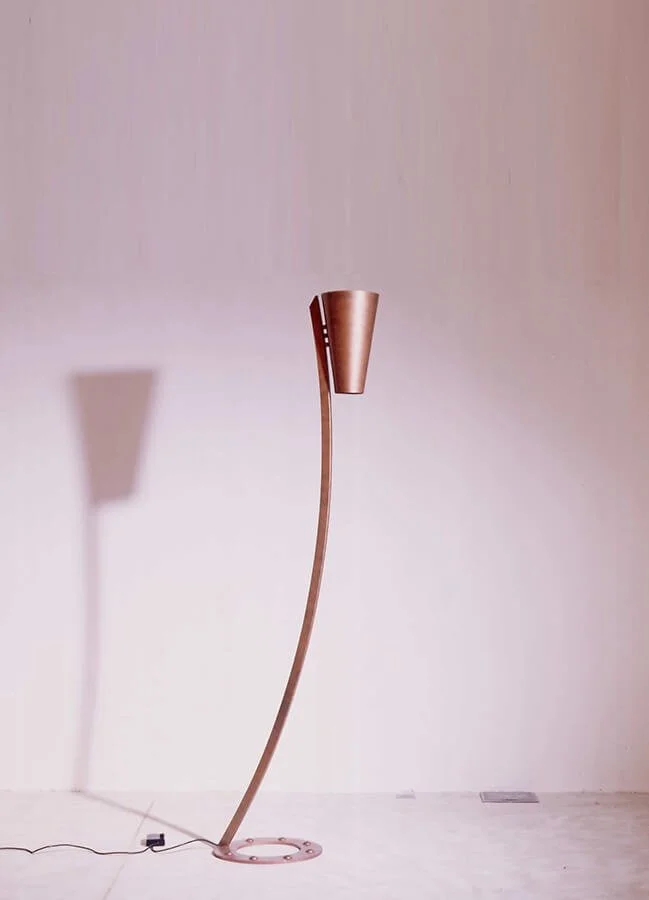 A tall, modern floor lamp with a curved stand and a copper-colored cone-shaped lampshade, casting a shadow on a plain light-colored wall.


