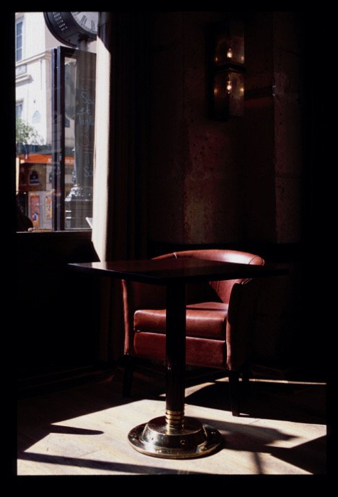 A dimly lit restaurant corner with a round wooden table and a pink upholstered armchair beside a window.


