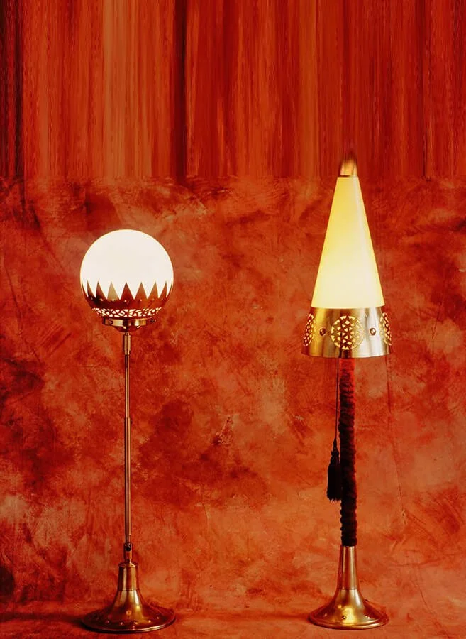 Two decorative floor lamps with gold bases and unique lampshades standing against a textured, reddish-brown wall.


