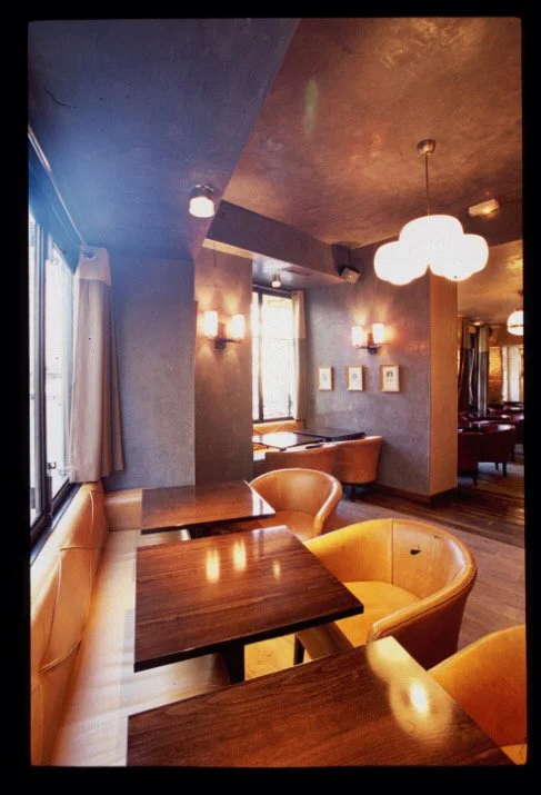 Cozy restaurant interior with wooden tables, mustard-colored chairs, and soft lighting from wall sconces and ceiling lamps.


