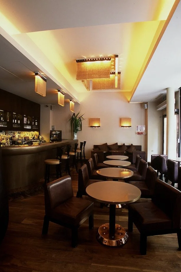 Interior of a modern lounge bar with round tables and leather chairs, a bar counter with bottles, soft warm lighting, and a cozy seating area with cushions.


