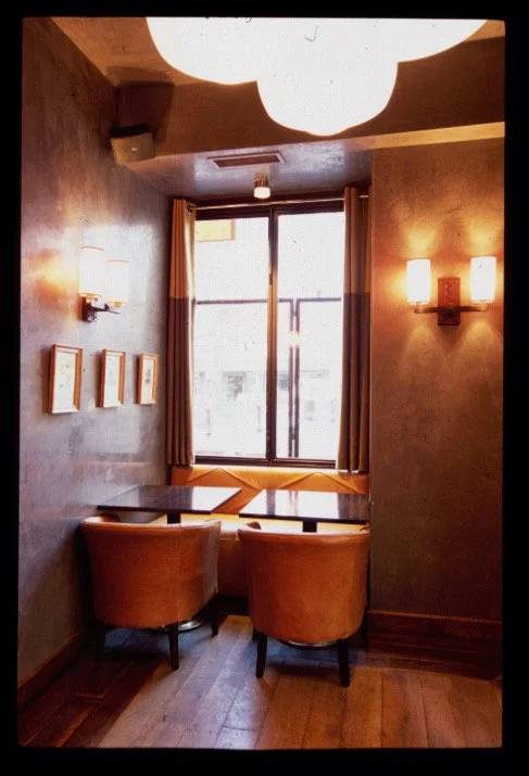 Cozy corner inside a cafe or restaurant with a window, a table, two orange chairs, and wall-mounted lights.


